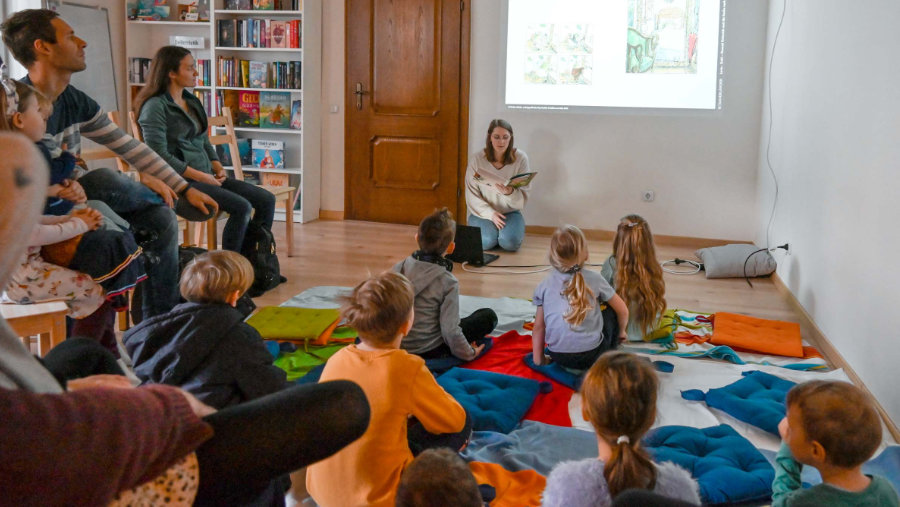 Ein Bilderbuchkino mit Konrad Kroeterich und der Suche nach der allerschönsten Umarmung in der erlesenen Bücherei Pottendorf mit vielen Kindern im Vordergrund, die gespannt Irina zuhören, wie sie die Geschichte vorliest.